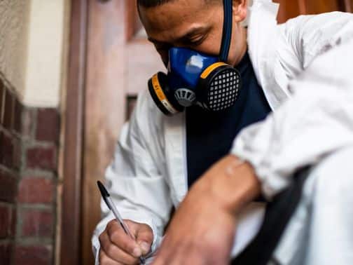 En person som bär andningsmask och skyddsdräkt skriver något på en urklipp medan han knäböjer bredvid en tegelvägg inomhus.
