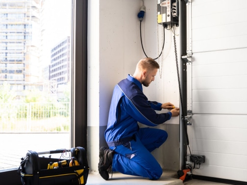 Tekniker som bär en blå uniform knäböjer vid en garagedörrsmekanism och gör justeringar med verktyg från en öppen verktygslåda i närheten.